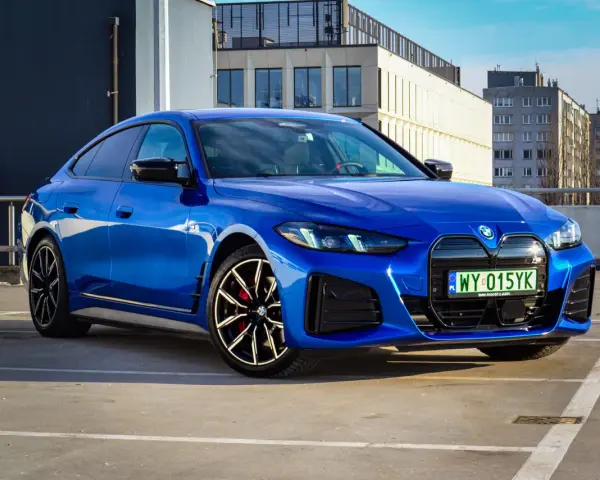 Niebieskie BMW i4 M50 zaparkowane na parkingu, z widokiem na budynki w tle.