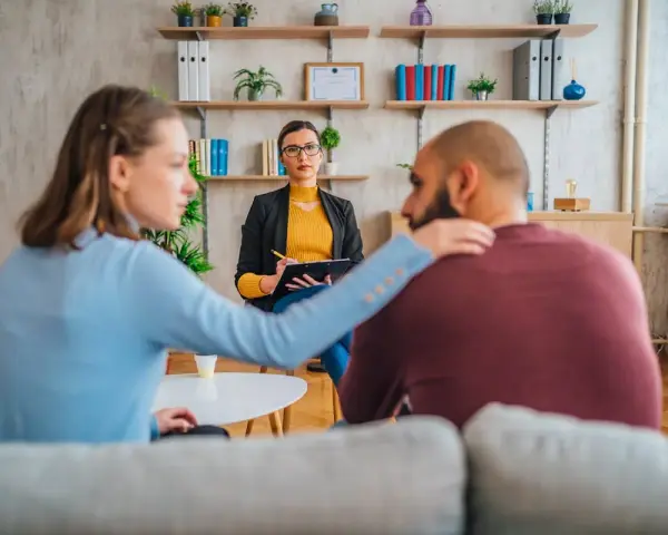 Un couple en consultation avec une psychologue conjugale. La femme pose une main réconfortante sur l'épaule de son partenaire.