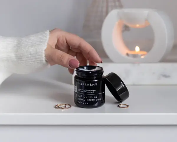 D'ALCHEMY - krem do twarzy, szyi i dekoltu. Dłoń sięga po słoiczek, w tle pali się świeca.