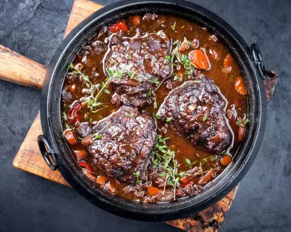 Carrilleras de ternera estofadas en salsa oscura con zanahorias y hierbas, servidas en una cazuela de hierro fundido sobre tabla de madera.