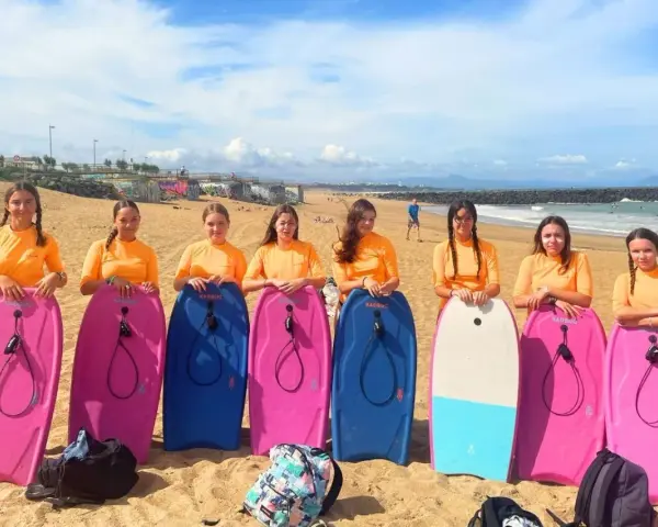 Huit jeunes filles en t-shirts orange, prêtes pour leur colonie à la mer, posent avec leurs bodyboards sur la plage.