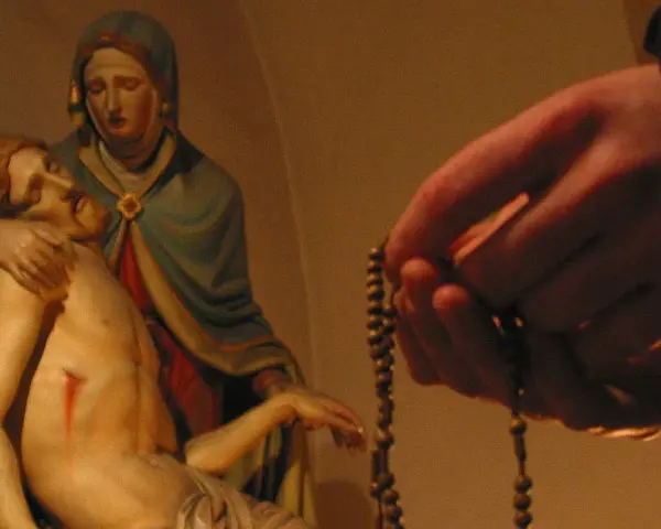 Eine Hand hält einen Rosenkranz, während die Pietà-Statue im Hintergrund zu sehen ist. Ein evangelischer Rosenkranz wird hier in Andacht gehalten.
