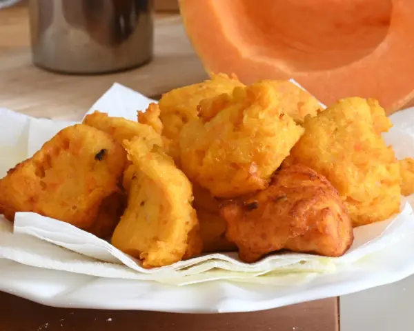 Frittelle di zucca dorate e croccanti, servite su carta assorbente, con una fetta di zucca sullo sfondo.