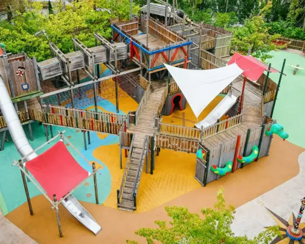 Ein riesiger, mehrstöckiger **Legoland Spielplatz** mit Rutschen, Kletternetzen und Holzbrücken, umgeben von grünen Bäumen.