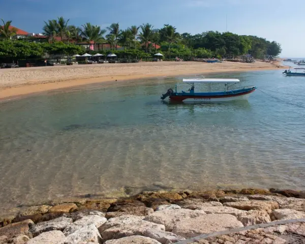 Spokojna zatoka w Sanur na Bali, z łodzią "Water Walk" kołyszącą się na turkusowej wodzie, plażą i palmami w tle.