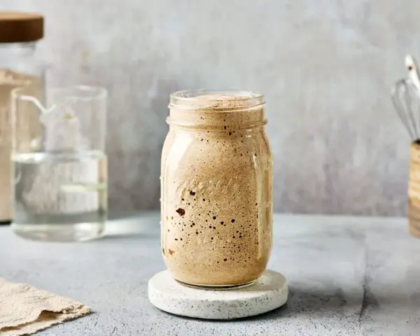 Ein Glas mit sprudelndem Sauerteig, angesetzt mit Dinkelmehl. Daneben ein Glas Wasser und ein Behälter mit Küchenutensilien.