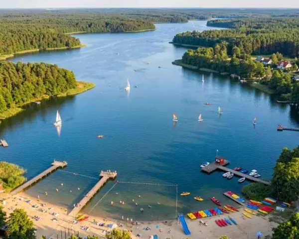 Letnie popołudnie nad jeziorem Kałębie. Żaglówki i kajaki na wodzie, plażowicze na piaszczystym brzegu, otoczonym zielonym lasem.
