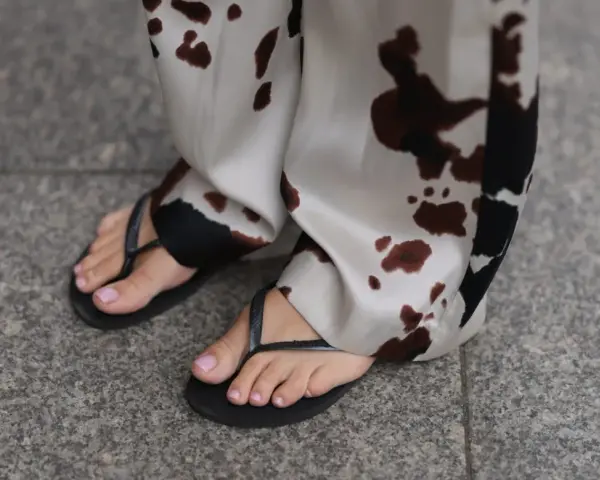 Delikatne, różowe paznokcie u stóp po zabiegu pedicure japoński, w czarnych klapkach, na tle spodni w cętki.