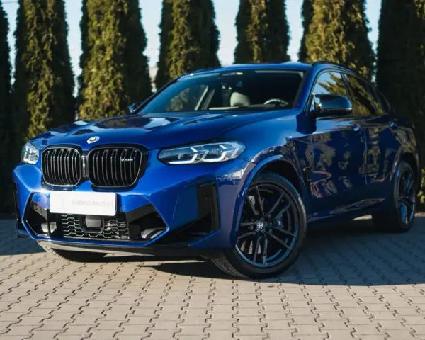 Błyszczący, niebieski suv BMW X4 M Competition zaparkowany przed żywopłotem.