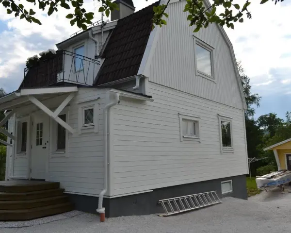 Vitt hus med ny fasad, stegar och material för byta fasad. En gul garagebyggnad syns i bakgrunden.