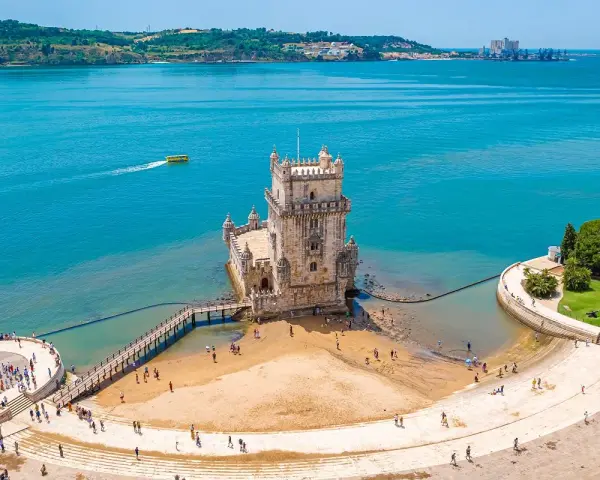 Wieża Belém, symbol kultury Portugalii, dumnie stoi nad wodą. Turyści spacerują po plaży i nabrzeżu.