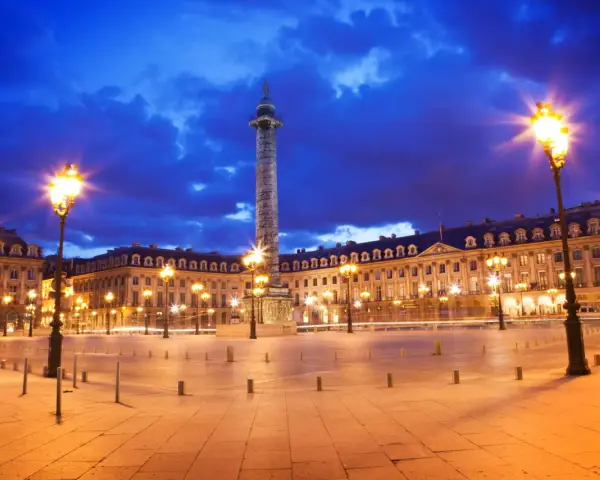La majestuosa plaza Vendôme se ilumina bajo un cielo nocturno azul intenso, con farolas doradas y la imponente columna en el centro.