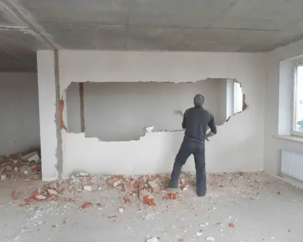 Mężczyzna z młotem wyburza ścianę nośną, tworząc w niej duży otwór. Wokół leżą gruzy.