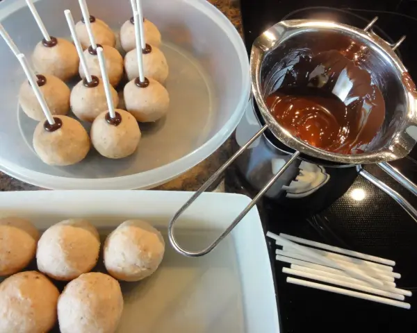 Biskuit-Cake-Pops warten auf ihre Schokoladenhülle. Geschmolzene Schokolade und Stäbchen liegen bereit.
