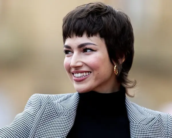 Uśmiechnięta kobieta z charakterystyczną fryzurą bixi, w czarnym golfie i wzorzystej marynarce.