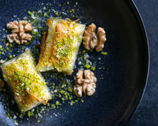 Zwei Stück Baklava, bestreut mit gehackten Pistazien und Nüssen, auf einem dunklen Teller. Ein wahrhaftiges Baklava Rezept Original.