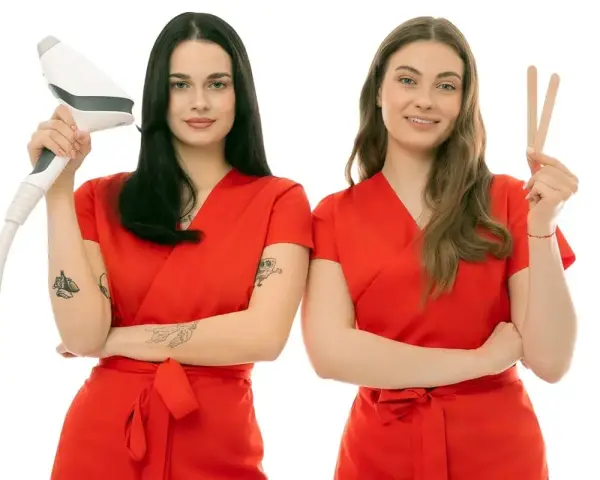 Dwie kosmetyczki w czerwonych uniformach. Jedna trzyma depilator, druga patyczki do wosku. Dowiedz się, ile kosztuje depilacja woskiem u kosmetyczki.