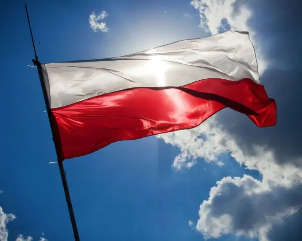 Biało-czerwona flaga Polski powiewa na tle błękitnego nieba z chmurami. Słońce przebija się przez materiał, podkreślając jego fakturę. Wymiary flagi polski są standardowe, proporcje zachowane.