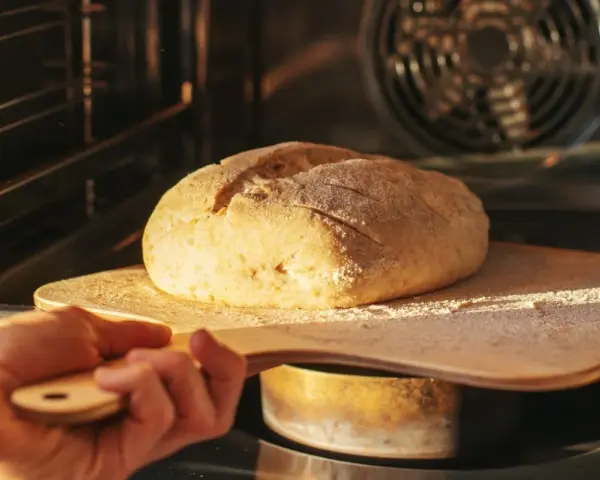 Ręka wsuwa drewnianą łopatą bochenek chleba do rozgrzanego piekarnika. To kulminacyjny moment wyrabiania ciasta na chleb.