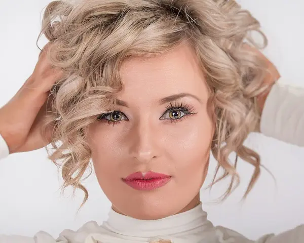 Blondynka z kręconymi włosami, z nonszalanckim gestem poprawiająca fryzurę. Jej oczy przyciągają uwagę, a usta są pomalowane na różowo.