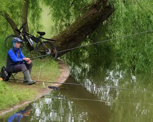 Wędkarstwo spławikowe na stawie. Mężczyzna w kasku i kamizelce siedzi na krześle, trzymając wędkę. Obok rower.