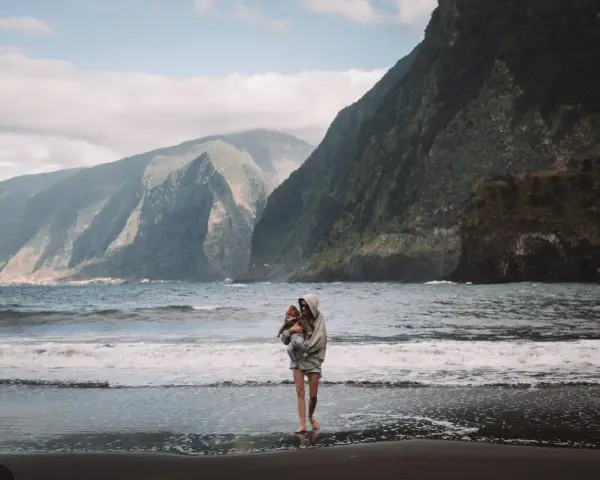Kobieta z dzieckiem na rękach na plaży, w tle majestatyczne góry i ocean. Niezapomniane madera widoki.