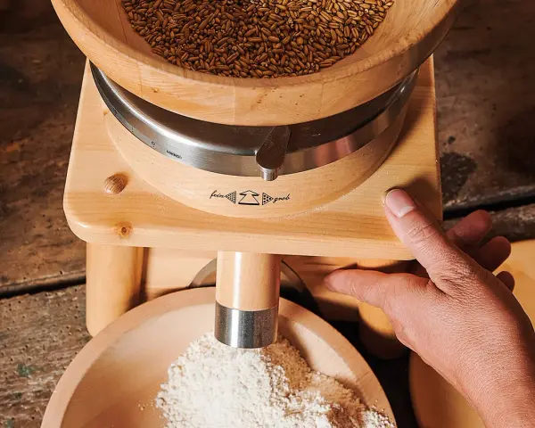 Holzgetreidemühle mit Getreide oben und frisch gemahlenem Mehl unten, Hand bedient die Mühle.