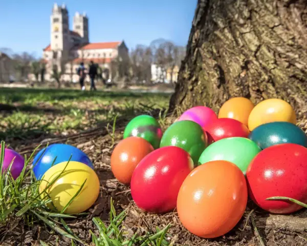 Bunte Ostereier liegen im Gras vor einem Baum. Im Hintergrund ist eine Kirche zu sehen. Die Suche nach den Eiern ist ein schöner Teil der Ostern Bedeutung.