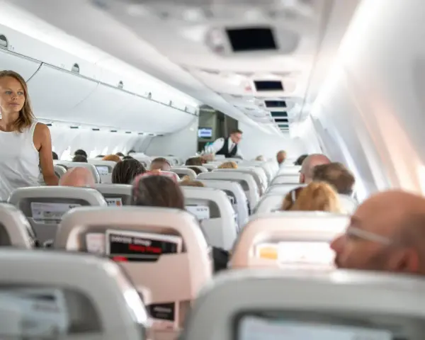 Widok wnętrza samolotu z pasażerami siedzącymi w rzędach i stewardesą.