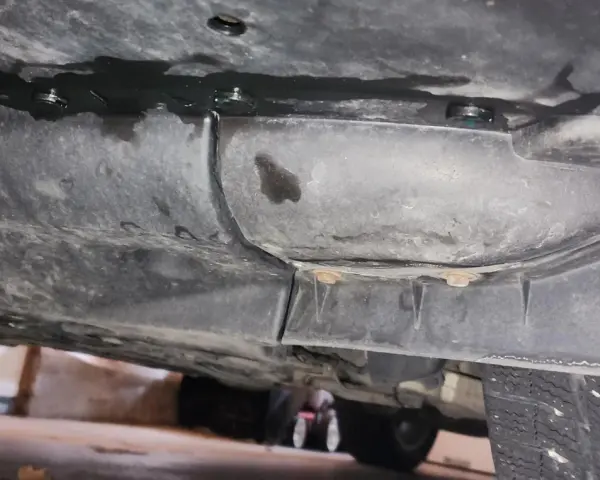 Underneath a car, dark fluid is leaking from a seam. A small puddle of blue fluid is visible, suggesting a coolant leak.