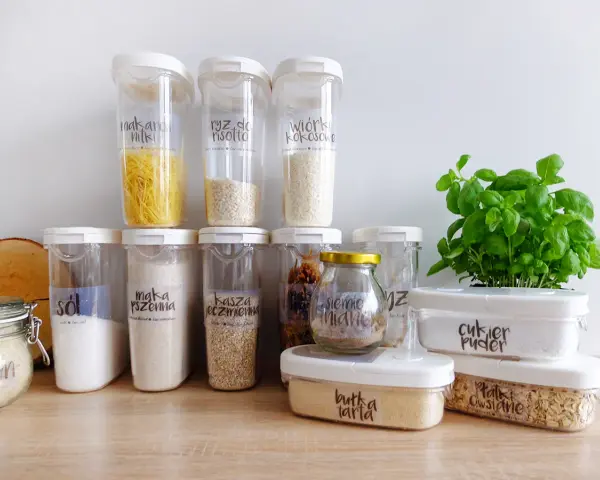 Pojemniki na żywność z etykietami pokazują, jak przechowywać pokrywki i produkty sypkie. Widać makaron, ryż, sól, mąkę, kaszę, cukier puder i płatki owsiane.