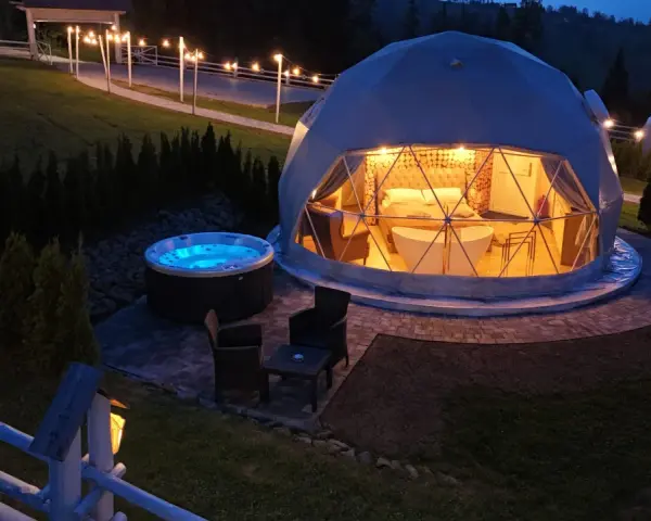 Nocny widok na luksusowy camping w Tatrach. Dwa kopułowe namioty, jeden z wanną i łóżkiem, drugi z oświetleniem. Obok jacuzzi i meble ogrodowe.
