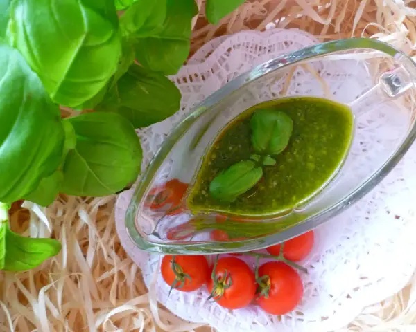 Aromatyczne pesto bazyliowe w szklanej sosjerce, ozdobione listkami bazylii, obok gałązki pomidorków koktajlowych.