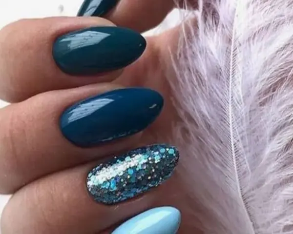 Unghie per l'inverno: unghie blu petrolio e azzurre con glitter scintillanti, accanto a una soffice piuma bianca.
