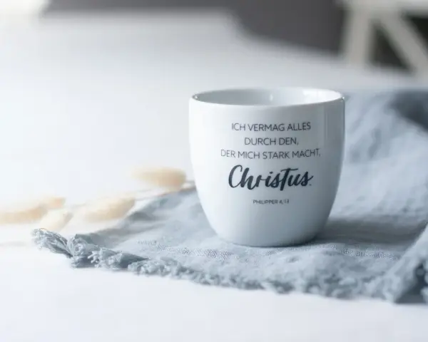 Weiße Tasse mit dem Schriftzug "Ich vermag alles durch den, der mich stark macht, Christus. Philipper 4,13" auf einem grauen Stoff.