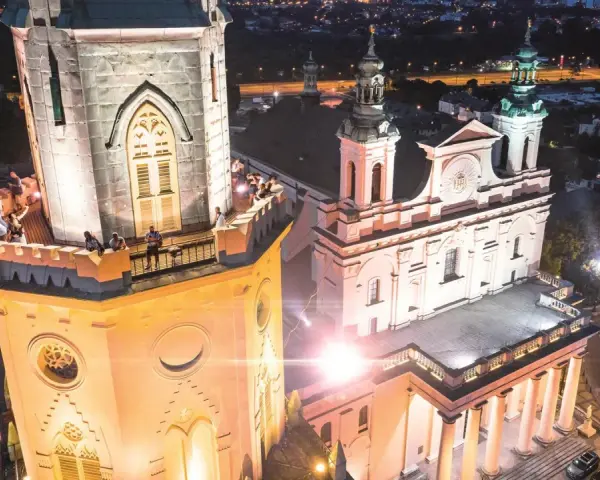 Wieża trynitarska nocą, z ludźmi na balkonie i oświetloną fasadą kościoła.