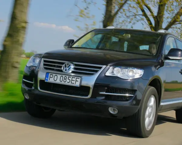Czarny VW Touareg 3.0 TDI w ruchu, z widocznymi wadami lakieru i drobnymi niedoskonałościami, na tle zielonych pól i drzew.