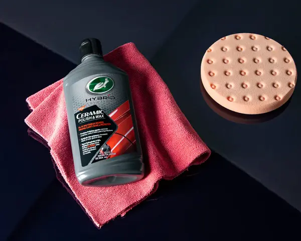 En flaska Turtle Wax Hybrid Solutions Ceramic Polish & Wax, en rosa mikrofiberduk och en polertrissa ligger på en mörkblå yta.