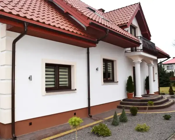Biała elewacja domu z brązowymi okiennicami i czerwonym dachem. Przed wejściem schody i zadbany ogród.