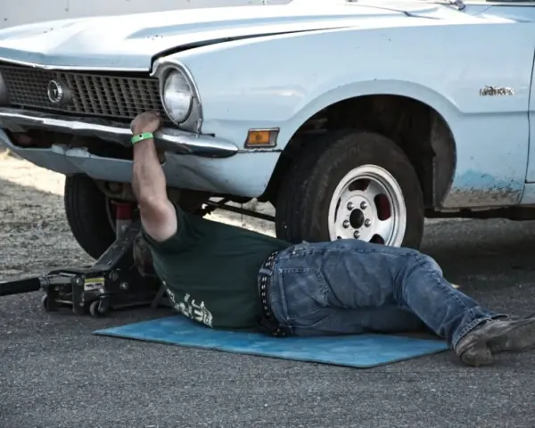 Un uomo lavora sotto un'auto d'epoca, forse per un intervento di **art 46 C.d.S.**, con un cric che la solleva.