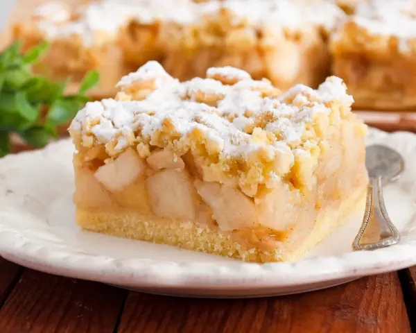 Kawałek szarlotki z kruszonką, posypany cukrem pudrem. Idealny na podwieczorek, według najsmaczniejsza szarlotka przepis.