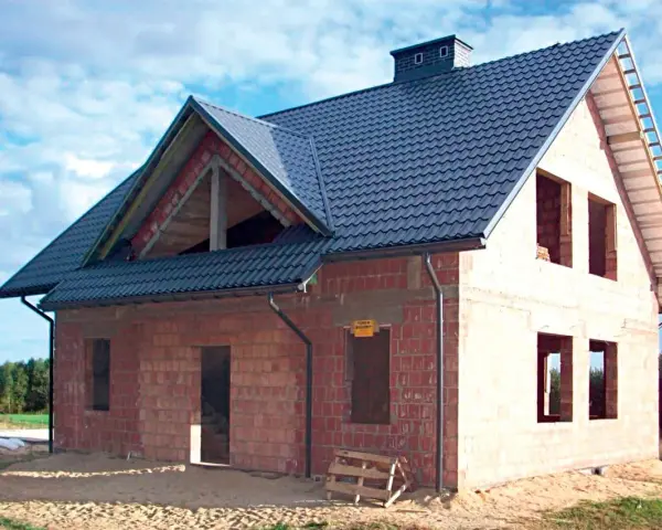 Budowa domu z cegły, widoczny dach betonowy skośny ocieplenie. Widać otwory okienne i drzwiowe, komin.