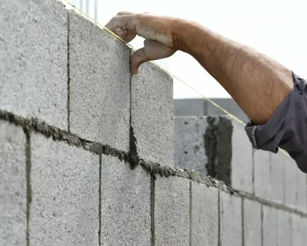 Main d'un maçon plaçant une parpaing pour monter un mur en parpaing de 2m. Le fil à plomb guide la pose.