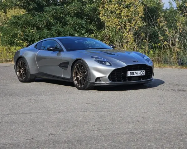 Srebrny Aston Martin Vanquish 2025 z brązowymi felgami na tle zieleni.