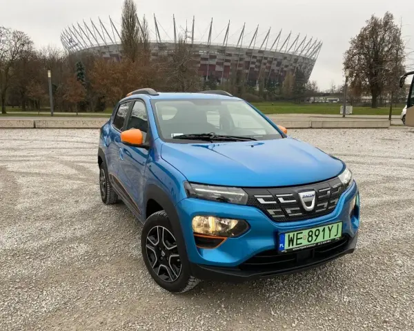 Niebieski Dacia Spring z pomarańczowymi akcentami na tle Stadionu Narodowego. Sprawdź atrakcyjną dacia spring cena!