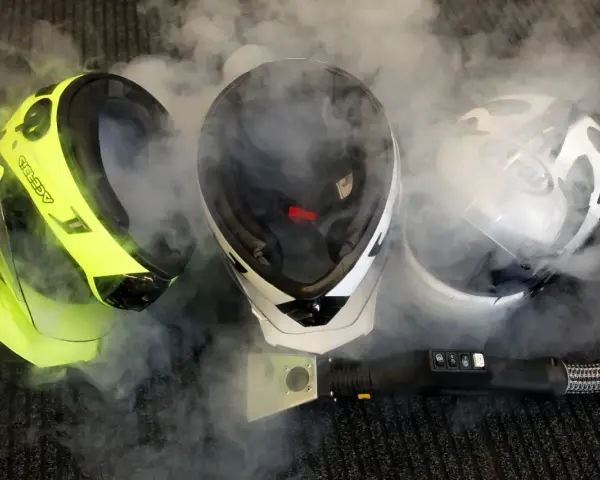 Pulizia del casco con vapore. Tre caschi, uno giallo fluo, uno bianco e nero, uno bianco e nero con visiera, circondati da fumo.