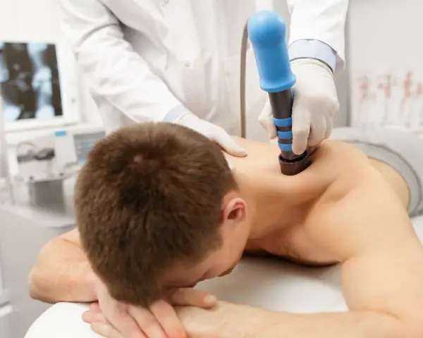 Arzt behandelt HWS-Syndrom mit Stoßwellentherapie. Ein Mann liegt auf dem Bauch, während ein Arzt mit einem blauen Gerät auf seinen Rücken drückt.