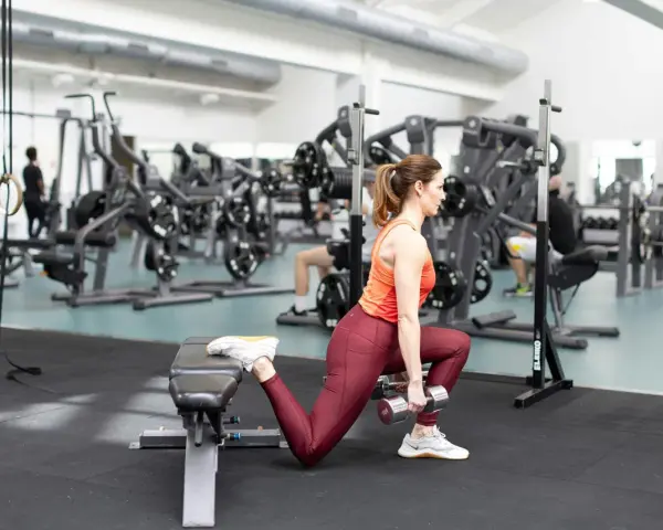 Kvinna utför bulgarian split squat med hantlar på gym.