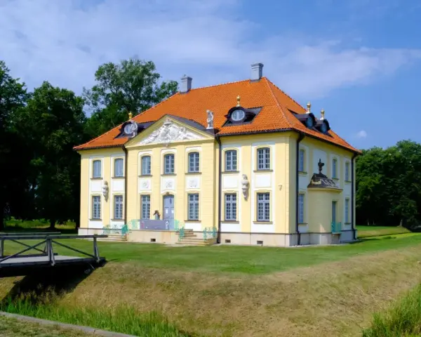 Pałac w Choroszczy, żółty, z pomarańczowym dachem, otoczony zielenią. Drewniany mostek prowadzi do posiadłości.