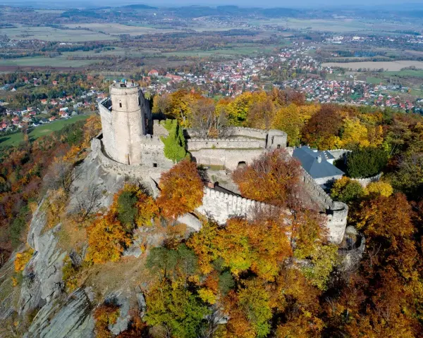 Zamek Chojnik na skalistym wzgórzu, otoczony jesiennymi barwami drzew. W oddali widać miasteczko i pola.